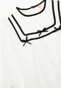 Vestido blanco para bebé con ribete negro, tres pequeños lazos negros en el pecho y escote redondo con cierre de botón en la parte posterior.