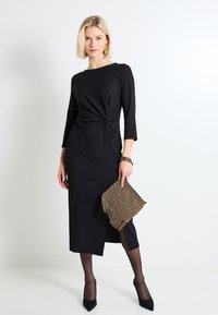 Vestido negro de manga larga con silueta ajustada y detalle delantero retorcido. Llevado con un bolso de mano dorado con flecos y tacones altos negros.