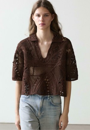 Ung kvinde med langt hår iført en brun hæklet kortærmet top kombineret med lyseblå jeans, stående foran en ensfarvet baggrund.