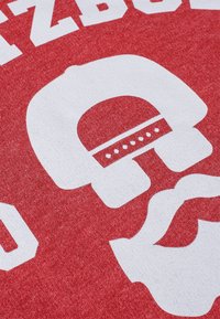 Graphique blanc d'un visage stylisé avec casque et moustache sur tissu rouge, avec des lettres blanches en bloc partiellement visibles au-dessus.