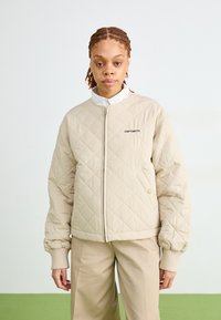 Giubbotto bomber trapuntato beige con zip frontale, polsini a costine e logo ricamato. Abbinato a pantaloni beige, sullo sfondo verde.