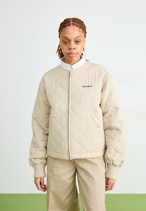 Giubbotto bomber trapuntato beige con zip frontale, polsini a costine e logo ricamato. Abbinato a pantaloni beige, sullo sfondo verde.