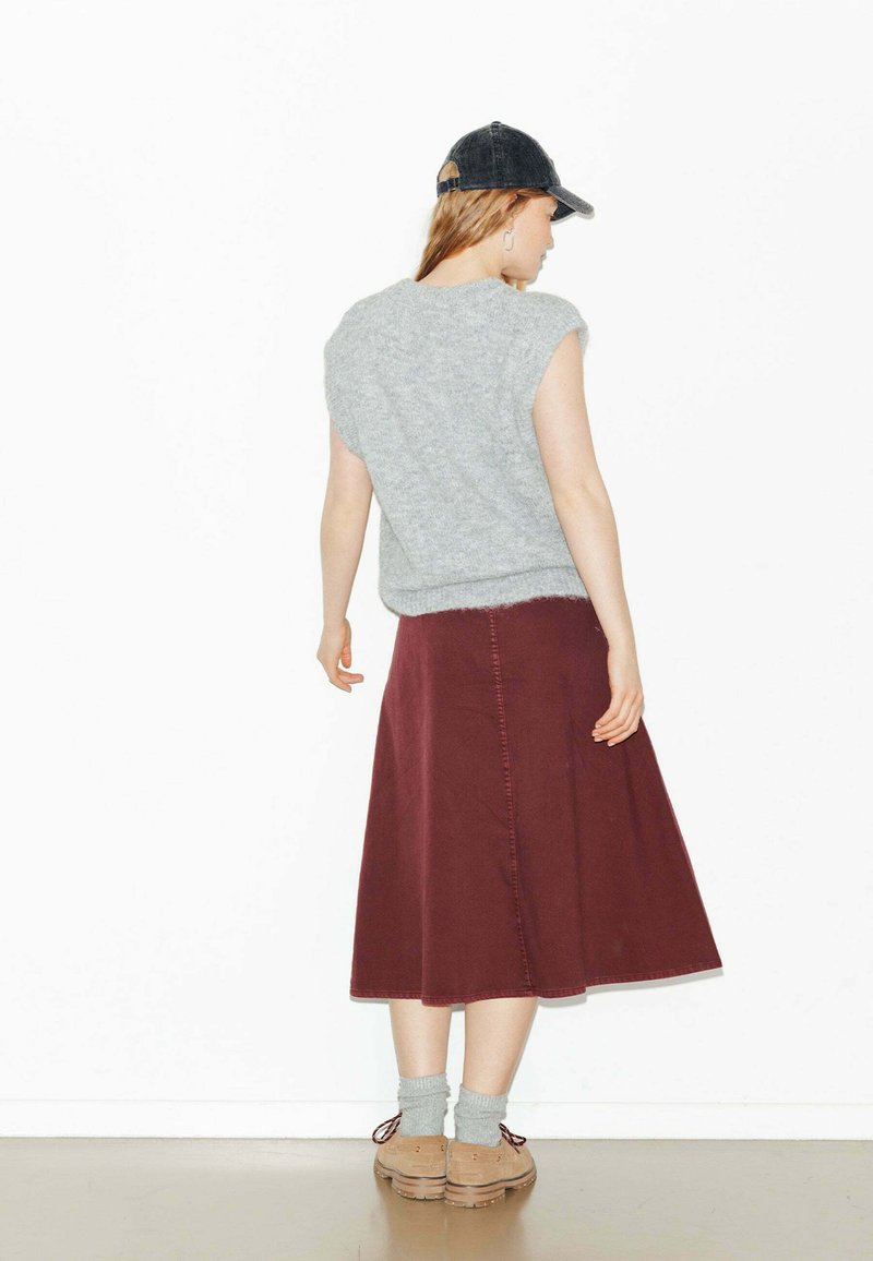 Pull en tricot gris à manches courtes associé à une longue jupe bordeaux. Le modèle porte des chaussettes grises et des chaussures beiges, coiffé d'un bonnet sombre.