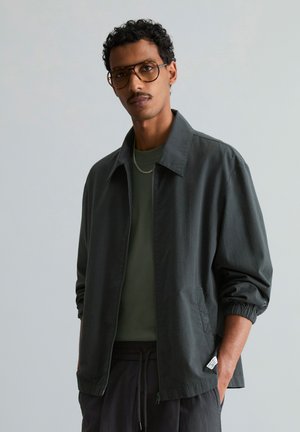 Man met krullend haar en snor, die een bruine bril draagt, donkergrijs vest met rits, groen shirt en een kettingketting tegen een effen achtergrond.