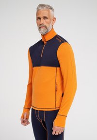 Fleece pullover med marineblå bryst og orange krop. Har en høj krave, halvdels lynlås og huller til tommelfingrene. Synede sømme og lynlås lomme.