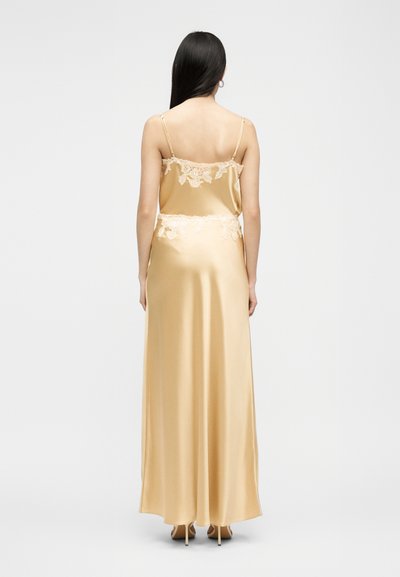 Γυναίκα με μακριά σκούρα μαλλιά, που φοράει χρυσό σατέν φόρεμα με δαντέλα στη πλάτη και τη μέση, στέκεται με την πλάτη γυρισμένη σε απλό φόντο.