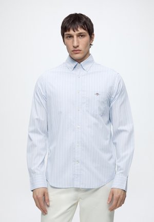 Uomo che indossa una camicia azzurro chiaro a maniche lunghe con sottili righe verticali e pantaloni bianchi, in piedi davanti a uno sfondo bianco semplice.