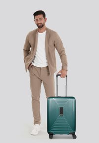 Valigia rigida in gradiente teal con design rigato, dettagli neri, manico telescopico e quattro ruote, posizionata accanto a un uomo in abbigliamento casual.