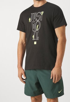Mężczyzna w czarnej koszulce Nike z napisem "JUST DO IT." i małymi żółtymi sylwetkami sportowców, w połączeniu z ciemnozielonymi szortami Nike.