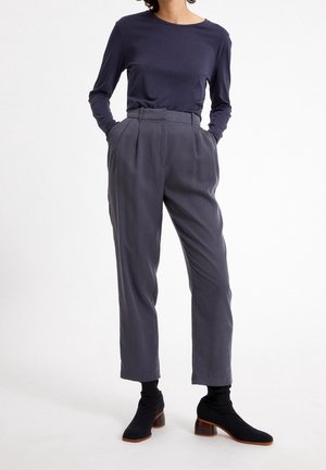 Osoba ubrana w granatową bluzkę z długim rękawem, szare plisowane spodnie z wysokim stanem oraz czarne botki na obcasie, stojąca z rękami w kieszeniach.