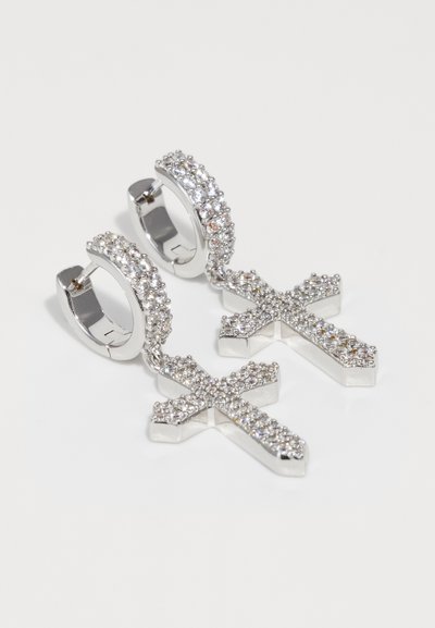 Boucles d'oreilles en argent en forme de créoles avec des pendentifs en croix suspendus, tous deux incrustés de petits cristaux clairs scintillants sur une surface blanche.