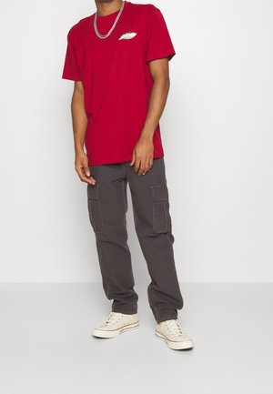 Rotes Baumwoll-T-Shirt mit einem weißen Grafikdruck auf der linken Brust, kombiniert mit grauen Cargo-Hosen und kremfarbigen Sneakern. Das Modell trägt eine silberne Halskette.