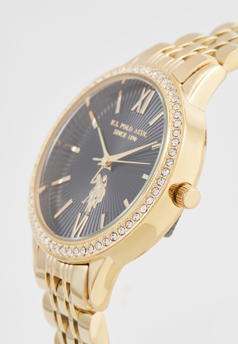 Goldfarbene Uhr mit einem runden schwarzen strukturierten Zifferblatt, Kristallakzenten um die Lünette und einem verlinkten Armband. Verfügt über römische Ziffern.