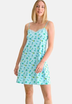 Femme portant une robe nuisette bleu clair à motifs floraux, avec des bretelles fines et une bordure en dentelle, se tenant devant un fond blanc uni.