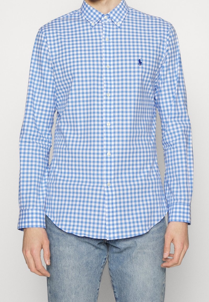 Mann trägt ein blau-weiß kariertes Hemd mit langen Ärmeln und eine hellblaue Jeans, steht vor einem weißen Hintergrund.