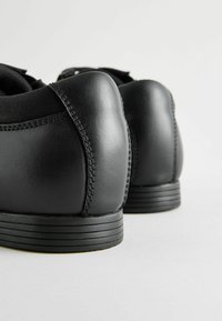 Zapatos de cuero negro con una textura suave, tacón redondeado y suela plana de goma. Presentan detalles de costura y un diseño minimalista.