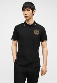 Νεαρός άνδρας που φοράει μαύρο πόλο μπλουζάκι με χρυσό περίγραμμα και κυκλικό λογότυπο "Versace Jeans Couture" στο στήθος, στέκεται με το ένα χέρι στην τσέπη.