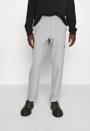 Mann trägt hellgraue, gerade geschnittene Hose mit Cargotaschen, schwarzes Langarmshirt und schwarze schnürschuhe vor einem schlichten Hintergrund.