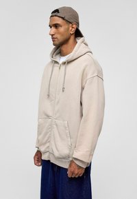 Beige zipper hoodie met trekkoord, voorzakken en geribde zoom. G gedragen met een wit shirt en losse blauwe denim broek. Licht stoffen textuur.