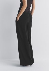 Pantalon large noir avec taille élastique et poches latérales. Texture lisse et coupe ample, conçu pour le confort et la liberté de mouvement.