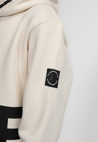 Felpa con cappuccio di colore crema, con un patchlogo nero sulla manica e una grande scritta nera sul davanti. Tessuto morbido e strutturato.