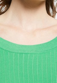 Haut vert en maille côtelée avec un large décolleté. Le tissu semble doux avec des rayures verticales, présentant une texture lisse et une coupe décontractée.