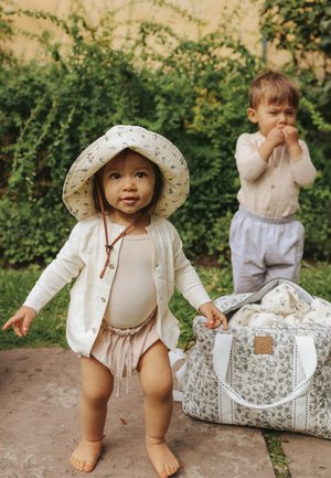 Bambino piccolo che indossa un cappello da sole e un cardigan, camminando a piedi nudi all'aperto, con un altro bambino in piedi vicino a una borsa con motivo floreale su un sentiero di pietra.