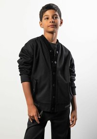 Czarna kurtka w stylu bomber z zapięciem na guziki, ściągaczami na rękawach i ukrytymi bocznymi kieszeniami. Miękki materiał o gładkiej fakturze.