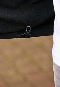 Jachetă din fleece negru, cu tiv ce prezintă un șnur cu un toggle negru și un cordon țesut. Textură netedă, strălucire subtilă, detalii de design minime.