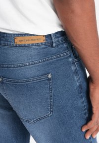 Vaqueros de mezclilla azul con una superficie texturizada, que presentan una etiqueta de cuero marrón que dice "ombre denim" y dos acentos de botón plateado en los bolsillos traseros.