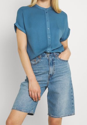 Femme portant une chemise bleue à manches courtes boutonnée, rentrée dans un short en jean bleu clair taille haute arrivant aux genoux.