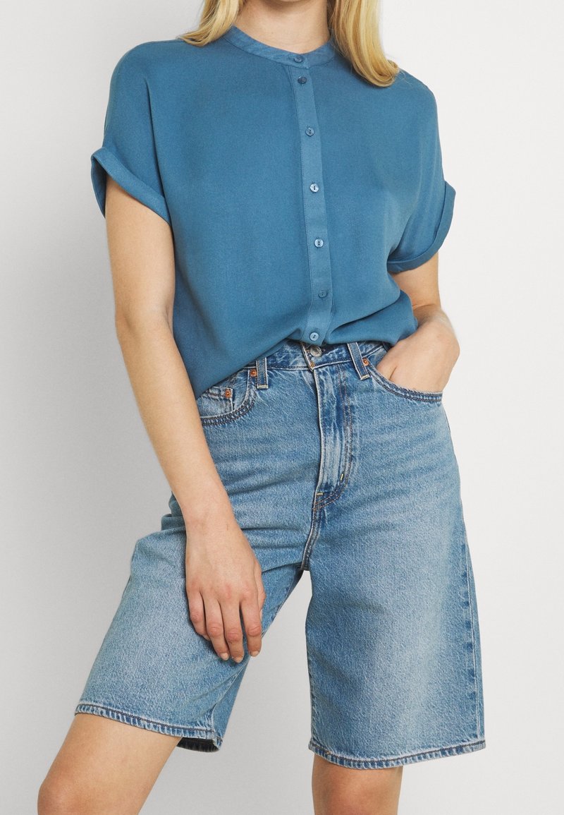 Femme portant une chemise bleue à manches courtes boutonnée, rentrée dans un short en jean bleu clair taille haute arrivant aux genoux.
