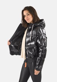 Femme portant une doudoune noire brillante avec capuche, la tenant ouverte pour montrer l'intérieur, associée à un haut beige et un pantalon en cuir noir.