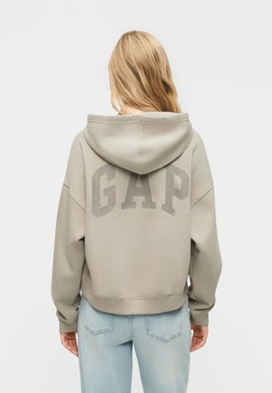 Ξανθιά γυναίκα που φοράει ανοιχτό μπεζ φούτερ GAP και ανοιχτό γαλάζιο τζιν, φαίνεται από πίσω με φόντο ένα απλό λευκό φόντο.