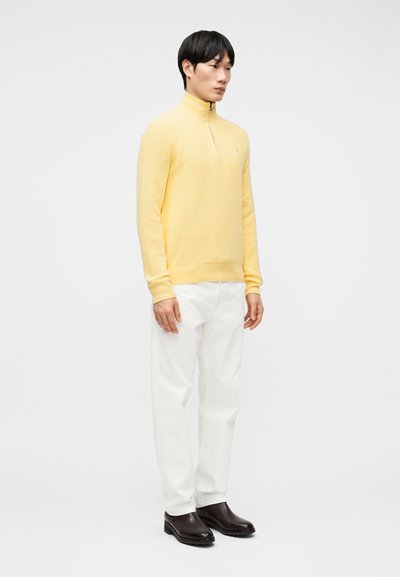 Άνδρας που στέκεται, φοράει κίτρινο πουλόβερ με φερμουάρ στο πάνω μέρος, λευκό παντελόνι και σκούρα καφέ δερμάτινα μποτάκια μπροστά σε απλό ανοιχτόχρωμο φόντο.