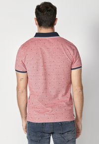 Červená polo tričko s námořnickým límečkem, krátkými rukávy a malými vzorovanými detaily. Materiál vypadá texturovaně. Nošeno s tmavými džínami.