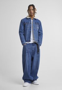 Jeans jakna z umetnim krznom okoli ovratnika, z sprednjimi žepi in vezenjem. Ohlapne jeans hlače, modre barve, zavarovane z črnim pasom.