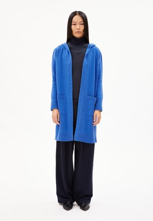 Langer, vorne offener Cardigan aus leuchtend blauer Strickware mit Kapuze, ausgestattet mit zwei vorderen Taschen und gerippten Bündchen. Kombiniert mit dunklen Hosen.