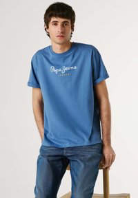 Jeune homme aux cheveux courts et foncés, portant un t-shirt bleu Pepe Jeans London et un jean bleu, assis sur un tabouret en bois devant un fond uni.