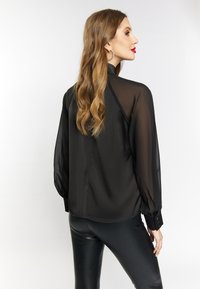 Sort gennemsigtig bluse med lange ærmer, høj krave og knaplukning ved håndleddene. Sørget for at matche med tætsiddende sorte bukser, der fremhæver en slank silhuet.