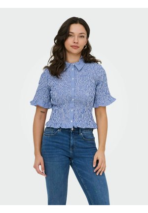 Femme aux cheveux ondulés foncés portant un chemisier à manches courtes à carreaux bleu et blanc et un jean bleu, debout devant un fond clair uni.