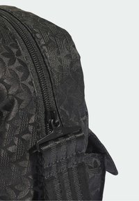 Zbliżenie czarnego plecaka z tkaniny z monogramem, czarnym zamkiem błyskawicznym oraz plecionym paskiem z plastikową klamrą.