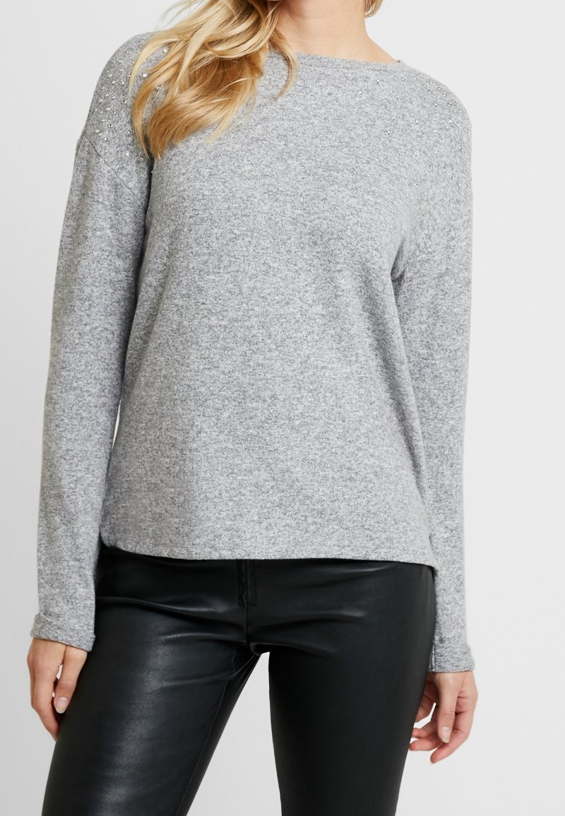 Femme portant un pull à manches longues gris clair avec de petites décorations sur les épaules et un pantalon en simili cuir noir.