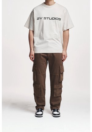 Camiseta de algodón beige con texto negro "2Y STUDIOS", combinada con pantalones cargo marrones que cuentan con múltiples bolsillos y un corte relajado. Zapatillas negras y blancas.