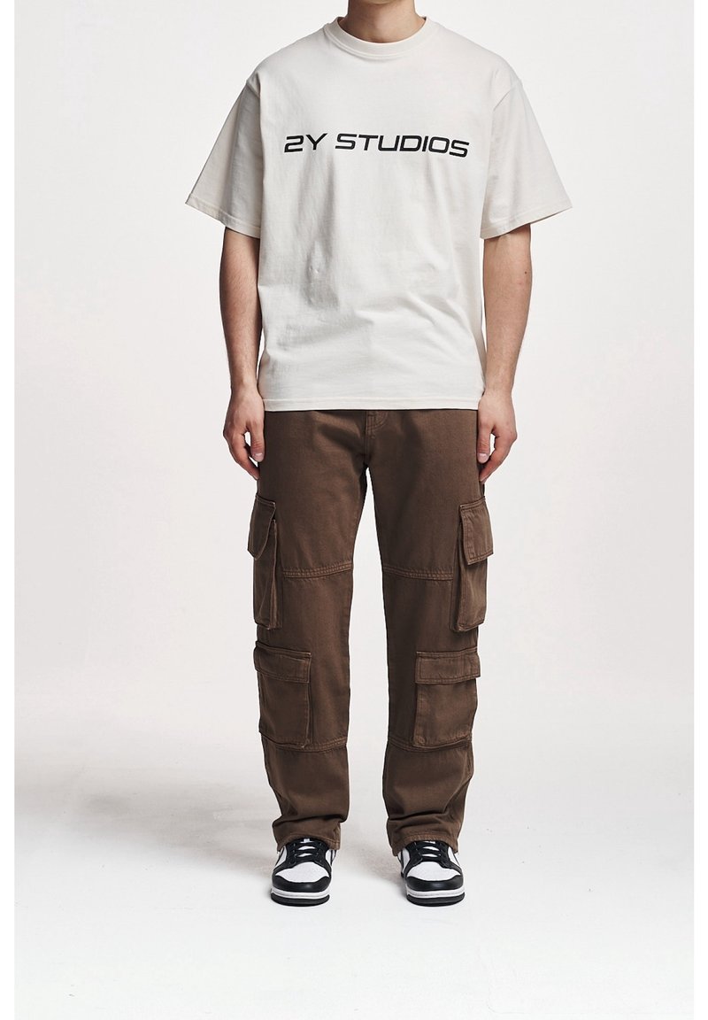 Beige bomullströja med svart text "2Y STUDIOS", kombinerad med bruna cargobyxor med flera fickor och en avslappnad passform. Svart-vita sneakers.