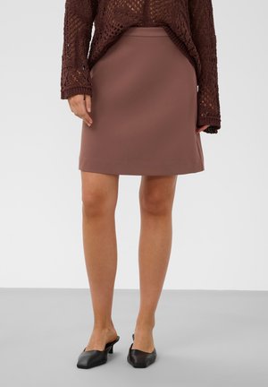 Femme portant une jupe marron longueur genou, un pull en maille marron et des chaussures noires plates à enfiler, debout sur un sol gris.