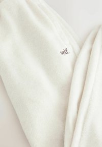 Valge fliiskangas, mille serva lähedale on väikeste tumedate tähtedega brodeeritud sõna "self".