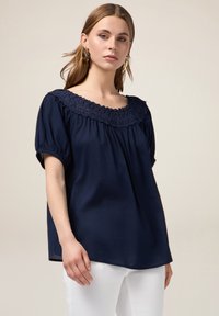 Blusa blu navy con scollatura in pizzo testurizzato, maniche a sbuffo corte e vestibilità ampia. Indossata con pantaloni bianchi, caratterizzati da un tessuto liscio.