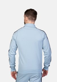 Helles blaues Sportjackett mit Kragen, das schwarze seitliche Streifen und lange Ärmel aufweist. Glatte Textur und figurbetontes Design.
