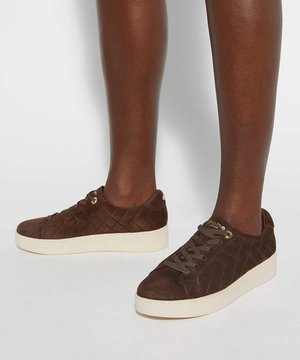 Baskets en daim marron avec motif matelassé et semelles épaisses crème portées sur des jambes à la peau foncée sur fond uni.